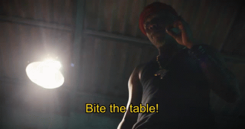 A Man Talking On A Cell Phone With The Words Bite The Table Written Below Him GIF