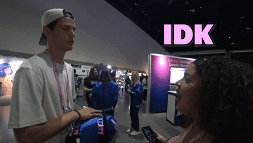 A Man Talking To A Woman In Front Of A Sign That Says Idk On It GIF