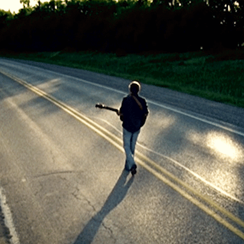 A Man Walking Down A Road With A Guitar GIF