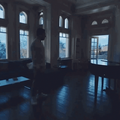 A Man Walking In A Dark Room With A Piano In The Background GIF