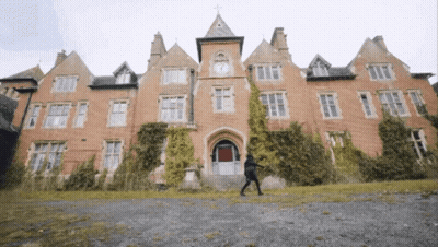 A Man Walking In Front Of A Large Brick Building With A Clock Tower GIF