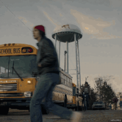 A Man Walking In Front Of A School Bus With A Water Tower In The Background GIF
