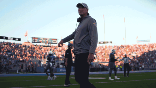 A Man Walking On A Football Field With A Scoreboard In The Background That Says ' Denver ' On It GIF