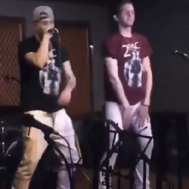 A Man Wearing A 2Pac Shirt Is Singing Into A Microphone While Another Man Stands Behind Him GIF