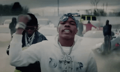A Man Wearing A Bandana And A Hoodie With The Word Baby On It Is Dancing In A Parking Lot . GIF