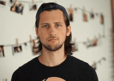A Man Wearing A Baseball Cap And A Black Shirt Stands In Front Of A Wall With Pictures Hanging On It GIF
