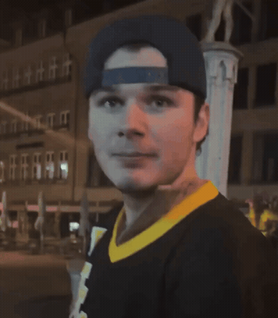 A Man Wearing A Baseball Cap And A Black Shirt With A Yellow Collar Looks At The Camera GIF