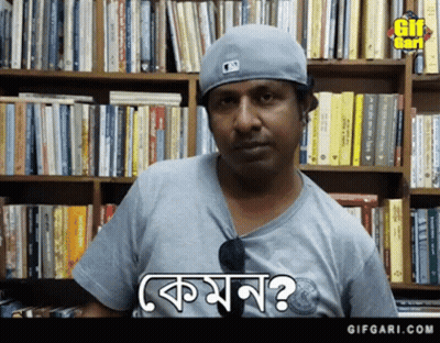 A Man Wearing A Baseball Cap And A Gray Shirt Is Standing In Front Of A Bookshelf And Says Komon GIF