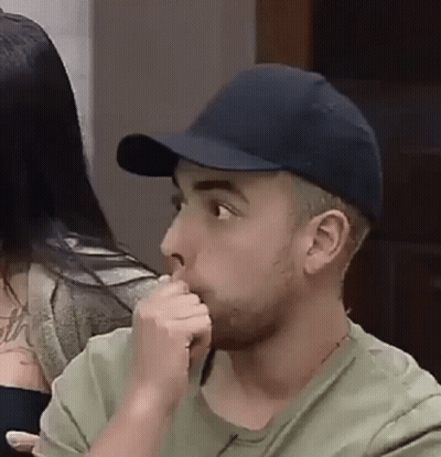 A Man Wearing A Baseball Cap And A Green Shirt Is Sitting At A Table With His Hand In His Mouth . GIF
