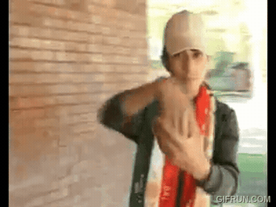 A Man Wearing A Baseball Cap And A Scarf Is Standing In Front Of A Brick Wall . GIF