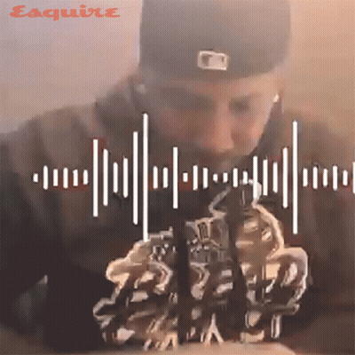 A Man Wearing A Baseball Cap And Ear Buds Looks Down At Something With Esquire Written On The Bottom Right GIF