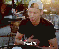 A Man Wearing A Baseball Cap Is Sitting At A Table And Says " What " GIF