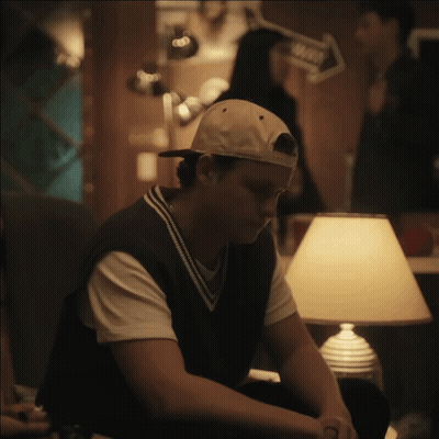 A Man Wearing A Baseball Cap Sits In Front Of A Lamp In A Dark Room GIF