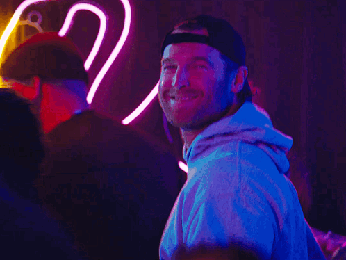 A Man Wearing A Baseball Cap Smiles In Front Of A Neon Sign GIF