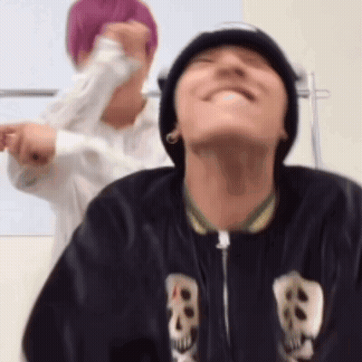 A Man Wearing A Beanie And A Jacket With Skulls On It Is Smiling While Another Man Stands Behind Him . GIF