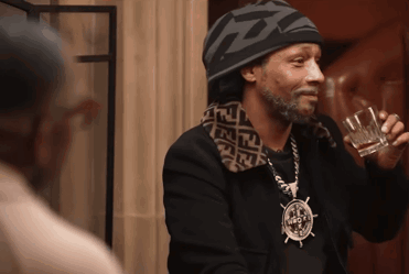 A Man Wearing A Beanie And A Necklace With A Compass On It Is Drinking A Glass Of Whiskey GIF