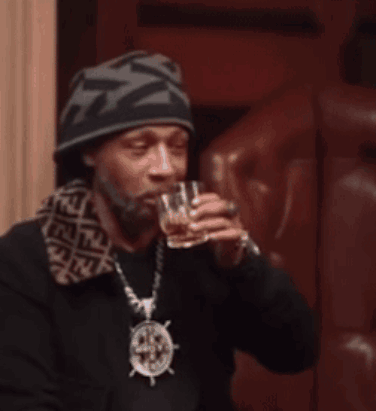 A Man Wearing A Beanie And A Necklace With A Steering Wheel On It Drinks From A Glass GIF