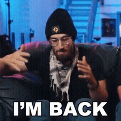 A Man Wearing A Beanie And Scarf Is Sitting On A Couch With The Words `` I 'M Back '' . GIF