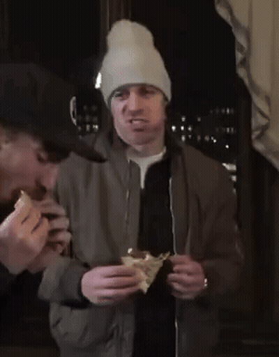 A Man Wearing A Beanie Is Eating A Sandwich While Another Man Watches GIF