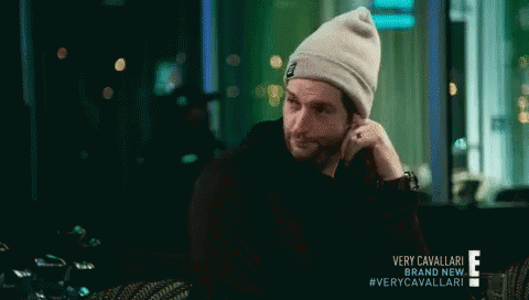 A Man Wearing A Beanie Is Sitting In A Dark Room With A Very Cavallari Brand New Advertisement Behind Him GIF