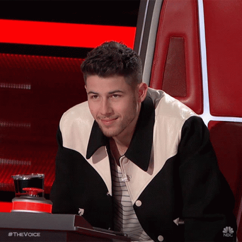 A Man Wearing A Black And White Jacket Is Sitting In A Chair With The Words #Thevoice On The Bottom GIF