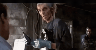 A Man Wearing A Black Apron And Gloves Is Talking To Another Man In A Dark Room . GIF