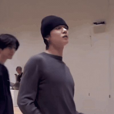 A Man Wearing A Black Beanie And A Gray Sweater Is Standing In A Room . GIF