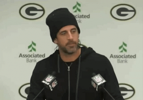 A Man Wearing A Black Beanie Stands In Front Of Two Microphones In Front Of A Wall With Green Bay Packers Logos On It GIF