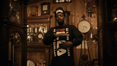 A Man Wearing A Black Fila Sweater Stands In Front Of A Wall Of Clocks GIF