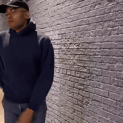 A Man Wearing A Black Hat And A Blue Hoodie Is Standing In Front Of A Brick Wall GIF