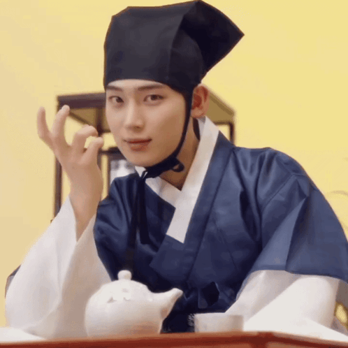 A Man Wearing A Black Hat And A Blue Robe Is Sitting At A Table GIF