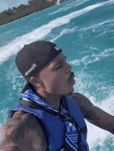 A Man Wearing A Black Hat And A Blue Vest Is Riding A Boat GIF