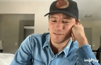 A Man Wearing A Black Hat And A Denim Shirt Is Sitting On A Couch . GIF