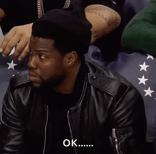 A Man Wearing A Black Hat And A Leather Jacket Is Sitting In A Stadium And Says Ok . GIF