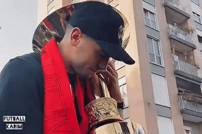 A Man Wearing A Black Hat And A Red Scarf Holds A Trophy GIF