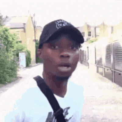 A Man Wearing A Black Hat And A White Shirt Is Standing On A Street . GIF
