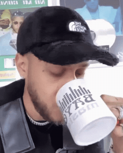 A Man Wearing A Black Hat Is Drinking From A White Mug That Says Bxa On It GIF