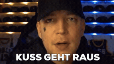 A Man Wearing A Black Hat Is Standing In Front Of A Wall Of Hats And Says Kiss Geht Raus . GIF