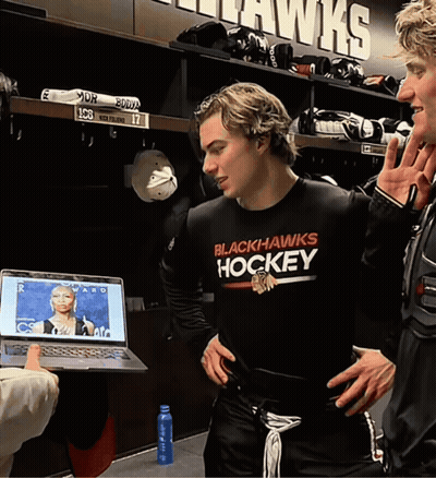 A Man Wearing A Black Hawks Hockey Shirt Stands In A Locker Room GIF