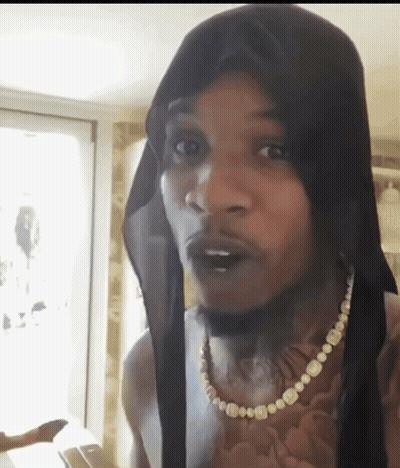 A Man Wearing A Black Head Scarf And A Pearl Necklace Looks At The Camera GIF