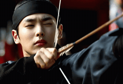 A Man Wearing A Black Headband Is Aiming A Bow And Arrow GIF