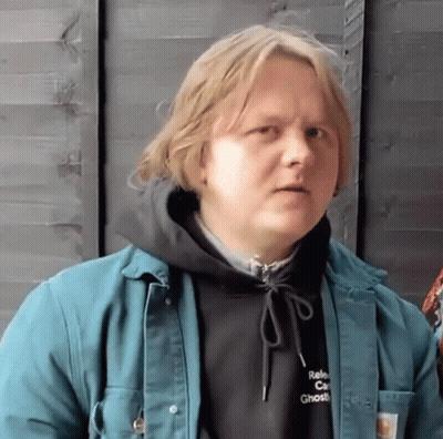 A Man Wearing A Black Hoodie And A Green Jacket Is Standing In Front Of A Wooden Fence . GIF