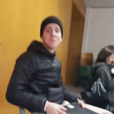 A Man Wearing A Black Jacket And A Black Hat Is Sitting In Front Of A Yellow Wall . GIF