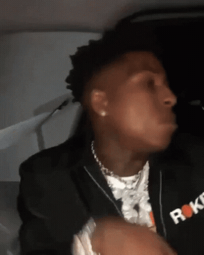 A Man Wearing A Black Jacket And A Necklace Is Sitting In A Car And Making A Funny Face . GIF
