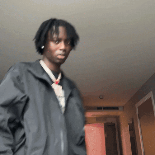 A Man Wearing A Black Jacket And A Necklace Is Standing In A Hallway GIF