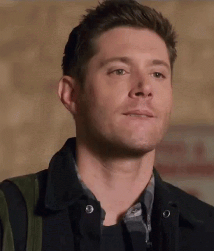 A Man Wearing A Black Jacket And A Plaid Shirt Is Smiling And Looking At The Camera . GIF