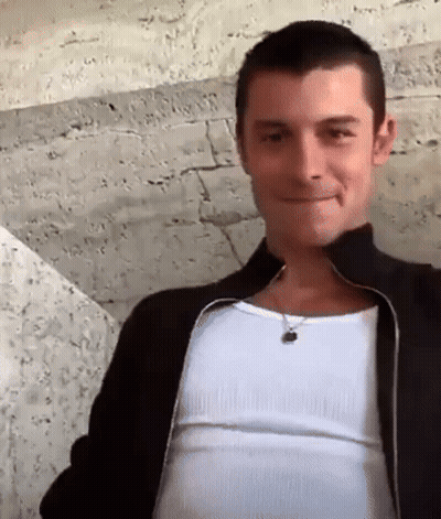 A Man Wearing A Black Jacket And A White Tank Top Smiles For The Camera . GIF