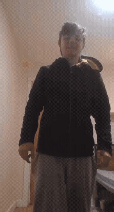 A Man Wearing A Black Jacket And Grey Pants Stands In A Hallway GIF