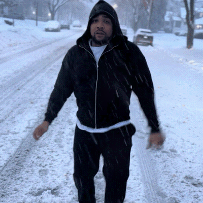A Man Wearing A Black Jacket With A Hood Is Walking In The Snow GIF