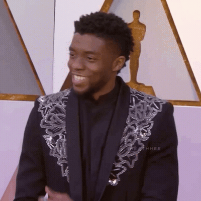 A Man Wearing A Black Jacket With Silver Sequins On The Sleeves Stands In Front Of A Statue Of An Oscar Winner GIF